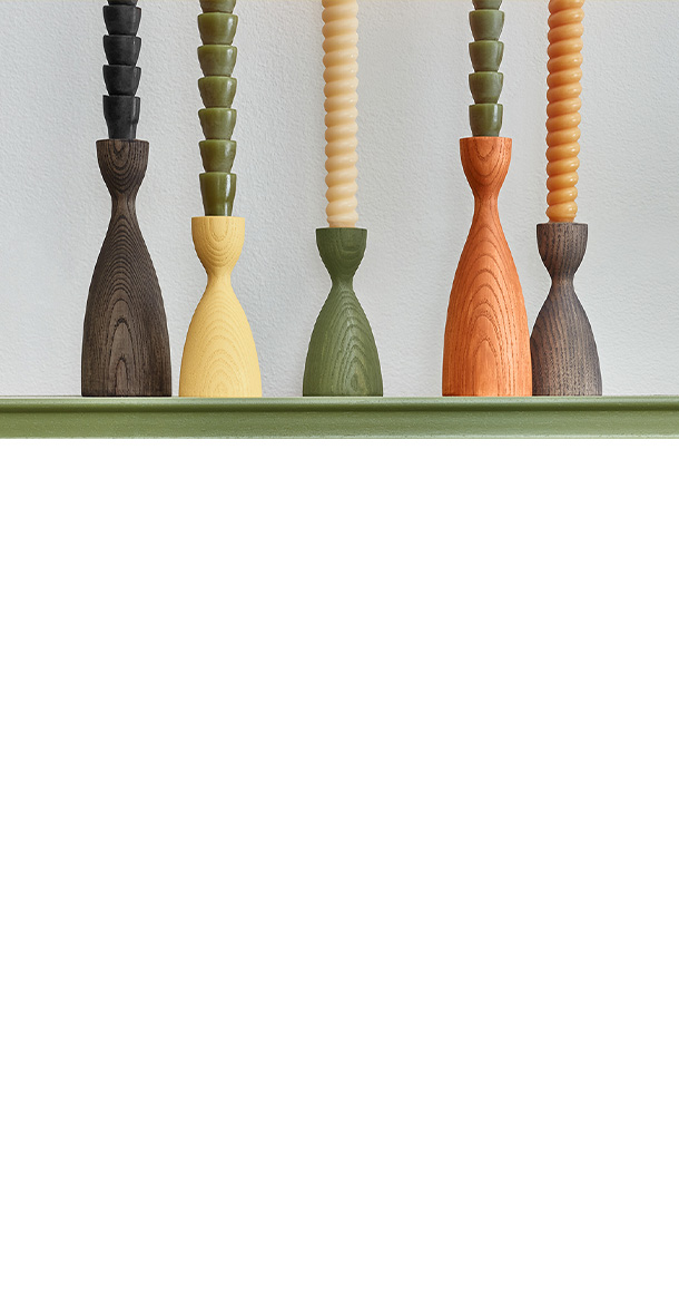 Five modern candlesticks with varied colors and wood-stains holders sit atop a buffet in a dining room, with a potted plant.